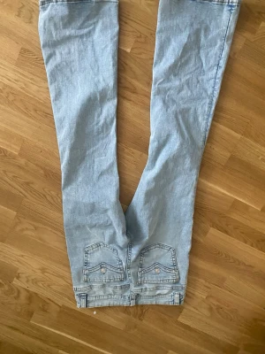 Ljusblå bootcut jeans med fickdetaljer - Säljer ett par ljusblå bootcut jeans med snygga fickor bak med lock och knapp. Jeansen har klassisk femficksdesign och är tillverkade i denim. Perfekta för en avslappnad och trendig look.
