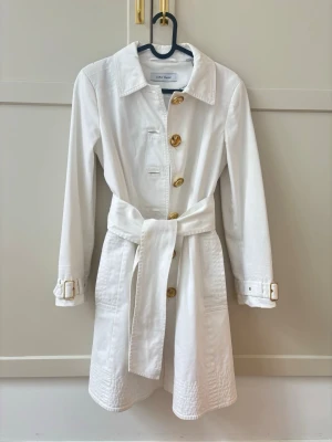 Vit trenchcoat Stl S från Zara Basic - Snygg vit trenchcoat från Zara Basic med knytskärp i midjan och guldiga knappar. Jackan har klassisk krage, långa ärmar med spännen vid ärmslut och stora fickor framtill. Perfekt för en stilren look under vår och sommar.