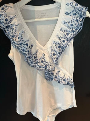 Vit body med broderier Intimissimi - Supersöt vit body från Intimissimi med blå blommiga broderier längs volangprydd omlottringning. 