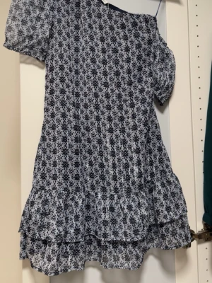 Blommig kortklänning med volang - Klänningen har puffiga korta ärmar och volangkant nedtill i lätt och luftigt material. Armen på höger sida kan tas bort. Helt ny klänning, oanvänd 💙 Skickar gärna fler bilder om det behövs :)