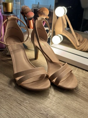 Beige höga sandaler från Graceland - Säljer ett par stilrena beige sandaler från Graceland med smala remmar och öppen tå. Skorna har en hög klack och justerbar ankelrem med spänne. Materialet är syntet och designen är enkel men elegant, perfekt för dig som gillar en clean look.