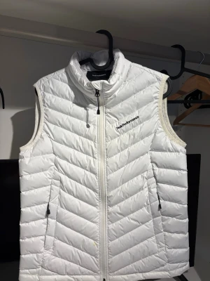 Vit dunväst från Peak Performance - Snygg vit dunväst från Peak Performance med dragkedja framtill och två fickor med dragkedja. Västen har en hög krage och stilrena, horisontella sömmar som ger en modern look. Perfekt för lager-på-lager och håller dig varm under kyliga dagar.