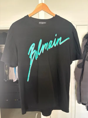 Svart Balmain t-shirt med turkost tryck - Cool svart t-shirt från Balmain med stort turkost logotyp-tryck framtill. Klassisk passform med rund hals och korta ärmar. Perfekt för dig som vill sticka ut med en statement-tee från ett exklusivt märke.