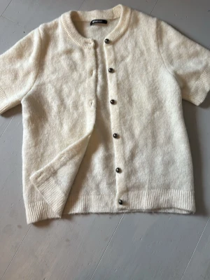 Krämvit kortärmad kofta - Krämvit stickad kofta från Gina Tricot, storlek S, med korta ärmar och rund hals. Koftan har ribbstickade muddar och knäpps med silvriga knappar framtill. Mjuk och fluffig känsla, extra knapp medföljer.