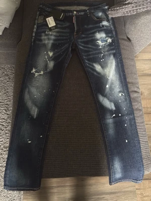 Mörkblå jeans med slitningar från dsq2 - Snygga mörkblå jeans från Belch med slitna detaljer och vita färgstänk för en trendig look. Jeansen har raka ben och klassisk femficksdesign. Perfekta för dig som gillar streetstyle och vill sticka ut lite extra.