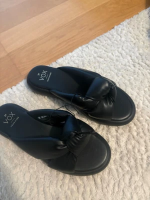 Svarta sandaler från Vox, storlek 39 - Säljer ett par svarta sandaler från Vox i storlek 39. De har en chunky, vadderad rem i veganläder och en mjuk fotbädd för extra komfort. Perfekta för en trendig och avslappnad look. Märkta med 'Vegan Shoes' på innersulan. Helt nya