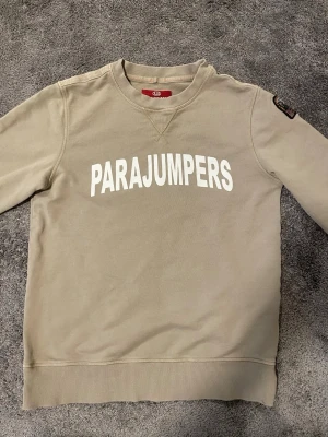Parajumpers tröja beige - Snygg beige sweatshirt från Parajumpers med stor vit logga på bröstet. Tröjan har rund halsringning, ribbade muddar och är gjord i mjukt fleecematerial. Perfekt för en avslappnad och trendig stil.