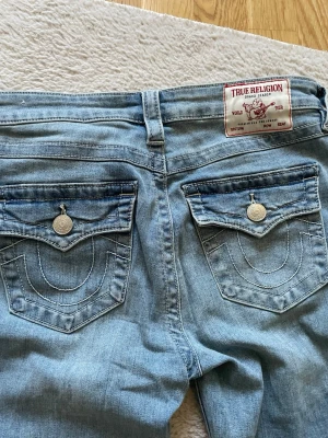 Ljusblå bootcut jeans True Religion - Säljer ett par ljusblå bootcut jeans från True Religion med ikoniska bakfickor och silverfärgade knappar. Jeansen har en klassisk femficksdesign och tydliga kontrastsömmar. Perfekta för dig som gillar en avslappnad och trendig look.