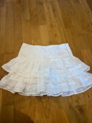 Vit volangkjol - Supersöt vit kortkjol med flera lager volanger och broderade detaljer. Kjolen har en luftig känsla och passar perfekt till varmare dagar. Den har en feminin  och lekfull stil med fina broderade mönster på tyget. Köpt från H&M men är tyvärr för liten och aldrig använd! Storlek Xs 