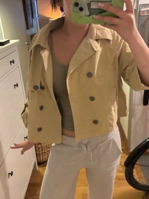 Trench coat - Snygg croppad jacka i beige med dubbelknäppning och stora knappar framtill. Jackan har krage och trekvartsärmar med knappdetalj vid ärmslut. Perfekt för dig som vill ha en trendig och avslappnad look.