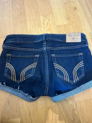 Hollister - Snygga mörkblå jeansshorts från Hollister med uppvikta ben och klassiska bakfickor med broderade detaljer. Shortsen har fem fickor, bälteshällor och slitningar framtill för en trendig look. Perfekta för sommaren! Lågmidjade och storlek 24