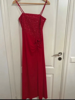 Röd långklänning med paljetter - Snygg röd långklänning med smala axelband och paljettprydd överdel. Klänningen har en dekorativ blomdetalj framtill och lätt drapering. Perfekt för dig som vill sticka ut med en elegant look.