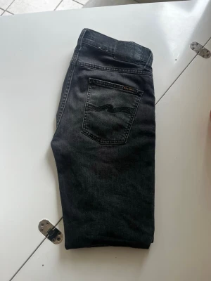 Svarta jeans från Nudie Jeans - Snygga svarta jeans från Nudie Jeans med klassisk femficksdesign och diskret logga på bakfickan. Jeansen har en smal passform och är tillverkade i stretchigt denimtyg för extra komfort. Perfekta för en stilren och avslappnad look. Storlek W29 L32