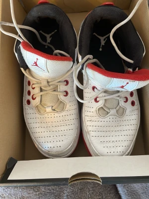 Vita Air Jordan sneakers med röd detalj - Säljer ett par vita Air Jordan sneakers med röda detaljer och svart insida. Skorna har klassisk perforerad tå, vita skosnören och den ikoniska Jumpman-loggan på plösen. Yttermaterialet är i läder och sulan är röd undertill. Perfekta för dig som gillar streetwear och sportig stil.