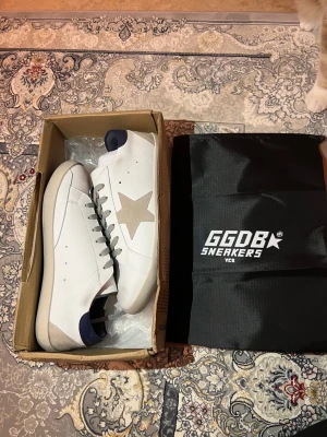 Golden Goose vita sneakers med stjärna - Golden Goose sneakers i vitt läder med beige mockadetaljer och ikonisk stjärna på sidan. Skorna har mörkblå hälflik, grå skosnören och klassisk låg siluett. Snygg kontrast mellan material och färger, perfekt för en trendig streetstyle.
