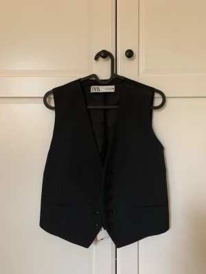 Svart kostymväst från Zara - Stilren svart kostymväst från Zara med klassisk passform och knäppning framtill. Fickdetaljer på båda sidor fram. Västen har en slät baksida i glansigt matrial med spänne i ryggen. Passform som en storlek S