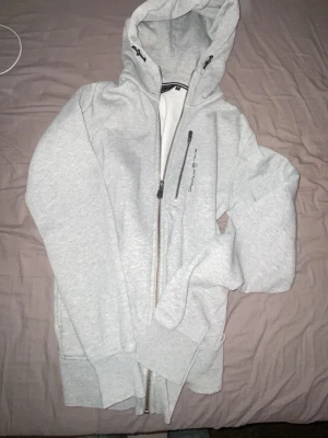 Ljusgrå hoodie från Sail Racing - Ljusgrå hoodie från Sail Racing med dragkedja framtill och justerbar huva med snören. Mjuk insida och ribbade muddar vid ärmar och nederkant. Diskret logga på bröstet och praktisk ficka med dragkedja. Perfekt för en avslappnad och sportig stil.