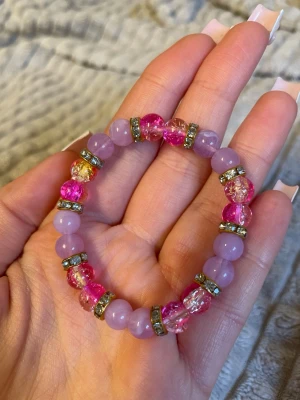 Lila och rosa pärlarmband - Säljer ett färgglatt armband med lila och rosa pärlor, varvat med glittriga mellanringar. Pärlorna är runda och sitter på en elastisk tråd, vilket gör armbandet lätt att ta på och av. Perfekt accessoar för att liva upp din outfit!