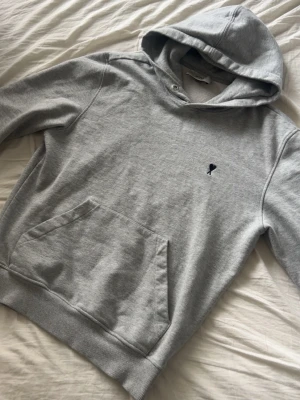 Grå hoodie från Ami Paris - Snygg grå hoodie från Ami Paris med klassisk känguruficka och liten svart Ami-logo broderad på bröstet. Hoodien har ribbade muddar vid ärmslut och nederkant samt en mjuk huva. Perfekt för en avslappnad och trendig stil.