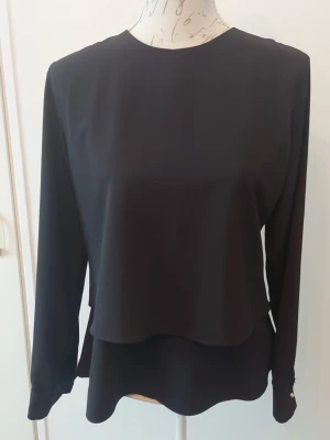 Svart långärmad blus med lager - Stilren svart blus med lång ärm och rund halsringning. Blusen har en snygg lager-på-lager-effekt framtill och knäppning med dragkedja i ryggen. Manschetter med knappdetalj ger en extra touch.