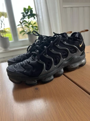 Nike Air VaporMax Plus svarta  - Svarta Nike Air VaporMax Plus sneakers med ikonisk vågig TPU-bur, mesh-överdel och synlig VaporMax-sula. Swoosh-logga i guld på hälen och dragflik bak. Grymt futuristisk look och riktigt sköna att gå i.