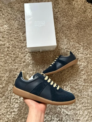 Mörkblå Maison Margiela sneakers - Snygga mörkblå sneakers från Maison Margiela med beige snörning och gummisula. Skorna har en mix av mocka och läderdetaljer samt en klassisk låg siluett. Perfekta för dig som gillar stilrena och tidlösa sneakers.