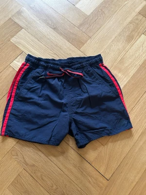 Adidas badbyxor  - Mörkblå badbyxor med röda ränder längs sidorna och matchande röd dragsko i midjan. Klassisk kort modell med elastisk midja och två sidofickor. Tillverkade i lätt och snabbtorkande syntetmaterial, perfekta för strandhäng.