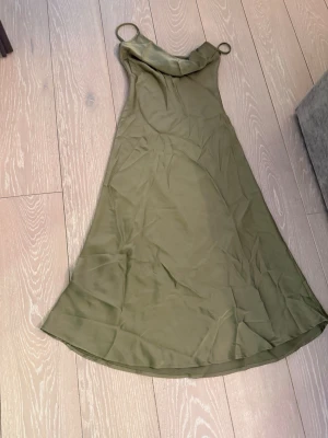 Olivgrön långklänning med smala band - Snygg olivgrön långklänning med smala axelband och lätt utsvängd passform. Klänningen har en enkel, stilren design och är tillverkad i ett mjukt, glansigt material som ger en elegant känsla.