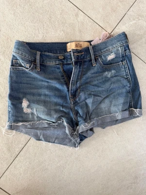 Blå jeansshorts med slitningar - Snygga blå jeansshorts från Hollister med slitna detaljer och fransig kant. Klassisk femficksmodell med orangea sömmar på bakfickorna. Perfekta för en avslappnad stil under varma dagar.