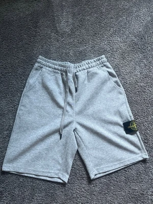 Grå shorts från Stone Island - Säljer ett par grå mjukisshorts från Stone Island med elastisk midja och snörning. Shortsen har två sidofickor och den klassiska Stone Island-loggan på vänster ben. Perfekta för chill dagar eller när du vill ha en avslappnad stil.
