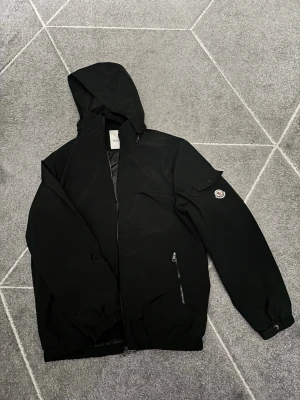 Svart vindjacka från Moncler - Stilren svart vindjacka från Moncler med huva och dragkedja framtill. Jackan har en ficka med lock och Moncler-logga på ärmen samt dragkedjeförsedd ficka på sidan. Tillverkad i ett lätt, vindtätt material som passar perfekt till streetwear.