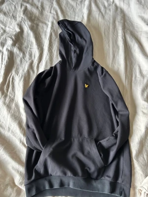 Svart hoodie från Lyle & Scott - Säljer en svart hoodie från Lyle & Scott med klassisk gul fågel-logga på bröstet. Hoodien har en stor huva och magficka framtill. Tillverkad i mjukt bomullsmaterial som är skönt att bära. Perfekt för en avslappnad och stilren look.