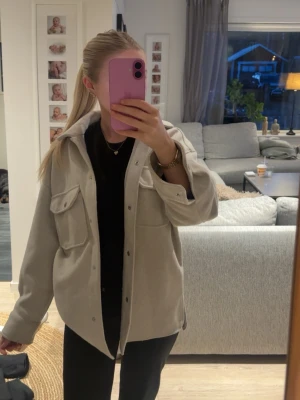 Beige/grå overshirt från Zara - Stilren beige/grå overshirt från Zara med två stora bröstfickor och tryckknappar framtill. Overshirten har en avslappnad passform och är gjord i ett mjukt, tjockare material som ger en snygg och trendig look. Perfekt att slänga över en hoodie eller t-shirt.