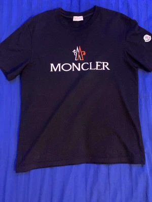 Svart Moncler t-shirt med logga - Snygg svart t-shirt från Moncler med klassisk logga i vitt och rött på bröstet samt patch på ärmen. T-shirten har rund hals och korta ärmar, tillverkad i mjuk bomull för en skön känsla. Perfekt för dig som vill ha en stilren och trendig look.
