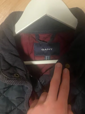 Mörkblå quiltad jacka från Gant - Snygg mörkblå quiltad jacka från Gant med ståkrage och detaljer i manchester. Jackan har knappar och dragkedja framtill samt justerbara spännen i sidorna. Perfekt för kyligare dagar och har en röd innerfoder som ger extra stil.