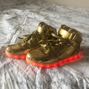 Guldiga sneakers med LED-ljus - Coola guldiga sneakers med snörning och LED-ljus i sulan som kan ändra färg. Perfekta för att sticka ut och lysa upp varje steg. Skorna har en högre modell med kardborreband för extra stöd. Parfekta till disco. Storlek 35 36 37 typ två lampor som lyser fel på vissa färger men inget som märks. Laddare till dom kan diskuteras.perfekt till barn som så på disco 🪩 