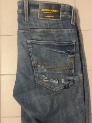 Blå slitna jeans från Jack & Jones - Säljer ett par blå jeans från Jack & Jones med slitningar och distressed detaljer. Jeansen har klassisk femficksmodell, raka ben och snygga kontrastsömmar på bakfickan. Perfekta för en avslappnad och trendig look.