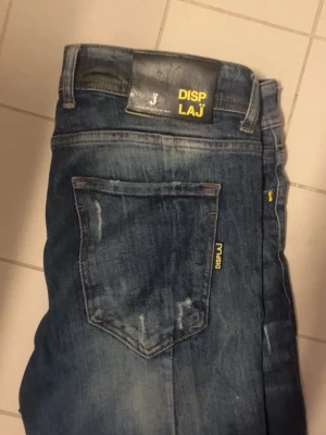 Blå jeans från DISPLAJ med slitningar - Säljer ett par blå jeans från DISPLAJ med snygga slitningar och tvättade detaljer. Klassisk femficksmodell med gul logotypdetalj på bakfickan och läderpatch i midjan. Perfekta för en avslappnad och trendig stil.