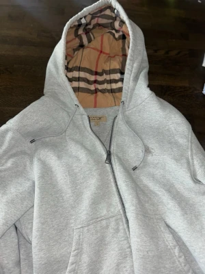 Grå zip hoodie från Burberry XL - Snygg grå zip hoodie från Burberry med klassiskt rutigt foder i huvan. Hoodien har dragsko i huvan, fickor framtill och är tillverkad i mjukt sweatshirtmaterial. Perfekt för en chill och stilren look.