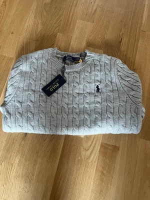 Ljusgrå kabelstickad tröja Polo Ralph Lauren - Snygg ljusgrå kabelstickad tröja från Polo Ralph Lauren med rund halsringning och klassisk broderad logga på bröstet. Tröjan har ribbade muddar och är tillverkad i mjukt material som passar perfekt till vardagslooken.