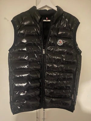 Svart dunväst från Moncler - Snygg svart dunväst från Moncler med glansig finish och klassisk logga på bröstet. Västen har dragkedja framtill och två sidofickor med dragkedja. S men passar M 