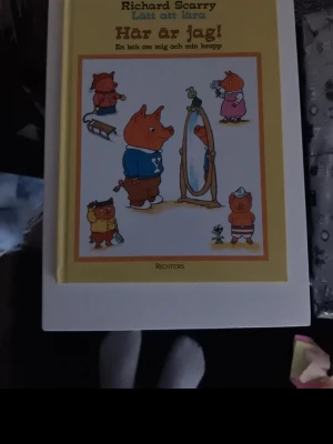 Här är jag! av Richard Scarry - Rolig och färgglad barnbok av Richard Scarry med titeln 'Här är jag!'. Boken har hårda pärmar i gult och vitt, och framsidan pryds av tecknade djurfigurer i olika kläder. Perfekt för dig som gillar klassiska bilderböcker.