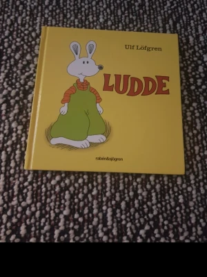 Ludde av Ulf Löfgren - En charmig barnbok med gul hård pärm, skriven av Ulf Löfgren. Omslaget pryds av Ludde, en vit kanin i gröna byxor och randig tröja. Perfekt för dig som gillar klassiska bilderböcker.