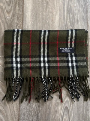 Grön rutig halsduk från Burberry - Snygg grön halsduk från Burberry i klassiskt rutigt mönster med inslag av svart, vitt och rött. Tillverkad i 100% kashmir och har fransar i ändarna. Perfekt accessoar för att lyfta din outfit med en lyxig touch.