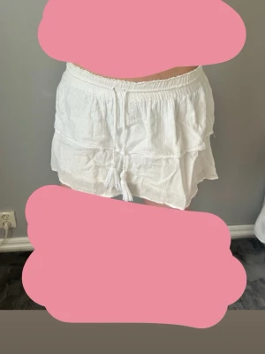 Vit kortkjol med volanger och snörning - Supersöt vit kortkjol med volanger och snörning i midjan. Kjolen har resår i midjan för skön passform och är gjord i ett lätt, luftigt material som passar perfekt till varma dagar. Enkel att matcha med olika toppar för en fräsch look.