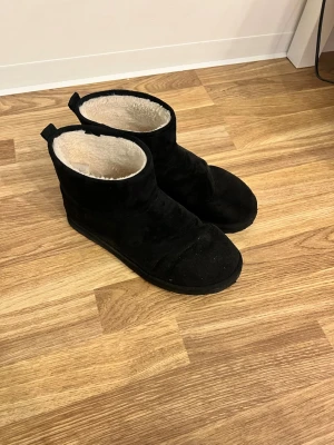 Svarta boots med foder - Säljer ett par svarta moon boots med mjukt vitt foder på insidan. Bootsens utsida är i mockaliknande material och de har en enkel, stilren design med rund tå och dragflik bak. Perfekta för kalla dagar när du vill hålla fötterna varma. Har fortfarande mycket kvar att ge!