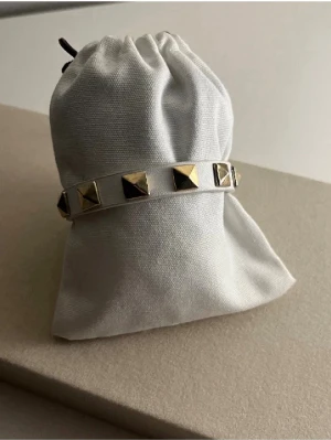 Vitt läderarmband med guldfärgade nitar - Snyggt vitt armband i läder med guldfärgade pyramidformade nitar runt om. Justerbart spänne i guldton ger en cool och edgy look. Perfekt accessoar för dig som vill sticka ut och addera lite attityd till din outfit.