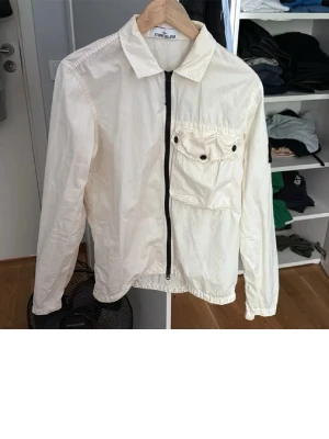 Vit vindjacka från Stone Island - Säljer en vit vindjacka från Stone Island med svart dragkedja framtill och en stor bröstficka med knappdetaljer. Jackan har klassisk krage och lång ärm, perfekt för dig som gillar stilrena och funktionella plagg.