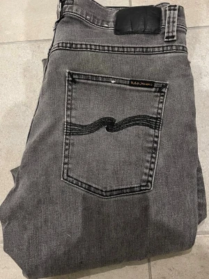 Grå jeans från Nudie Jeans - Snygga grå jeans från Nudie Jeans med raka ben och klassisk femficksdesign. Jeansen har svarta detaljer på bakfickan och svart patch i midjan. Tillverkade i denim med en cool tvättad look och uppklippta där ner. Storlek 32/31