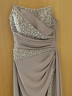 Lång beige klänning med pärlor - Elegant långklänning i beige med en axelbandslös design och draperad front. Klänningen är dekorerad med mängder av pärlor upptill och längs midjan, vilket ger en lyxig känsla. Tillverkad i ett mjukt, flytande tyg som faller snyggt.